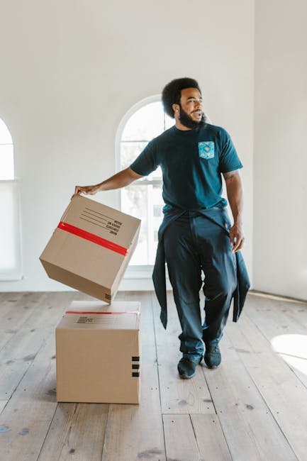 A man with dark curly hair and a beard, dressed in a navy blue t-shirt and matching trousers, is holding a cardboard box wrapped in brown packing tape with a red stripe. He is standing inside a spacious, well-lit room with white walls, wooden flooring, and large arched windows allowing natural light to brighten the area. The man appears to be preparing for a home relocation, with a second cardboard box placed on the floor nearby, both boxes indicating staff involvement in packing or loading. The environment suggests a professional moving process, supported by the presence of packing materials and the open space suitable for loading furniture and boxes onto a moving vehicle. The light and shadows highlight the clarity of the scene, emphasizing the careful handling involved in furniture transport and packing during house removals, as undertaken by Movers Hounslow.