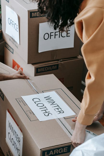 Close-up view of a professional moving process with a focus on packing and loading office items, featuring a person’s hands securing large cardboard boxes labeled 'WINTER CLOTHING' on a flat surface. Behind, multiple medium-sized cardboard boxes with white labels marked 'OFFICE' are stacked inside a room, some with plastic wrapping or packing tape. The boxes are arranged on the floor near a doorway or entry area, with natural or artificial lighting highlighting the details of the packaging materials. Tools such as a lifting strap and a trolley may be partially visible, indicating a systematic approach to furniture and goods transport. This scene is representative of home or office relocation activities managed by Movers Hounslow as part of their removals services, demonstrating the careful packing, organization, and logistics involved in a furniture transport or moving operation.