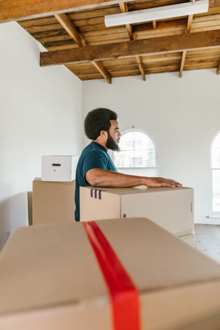 Close-up view of a professional moving process with a focus on packing and loading office items, featuring a person’s hands securing large cardboard boxes labeled 'WINTER CLOTHING' on a flat surface. Behind, multiple medium-sized cardboard boxes with white labels marked 'OFFICE' are stacked inside a room, some with plastic wrapping or packing tape. The boxes are arranged on the floor near a doorway or entry area, with natural or artificial lighting highlighting the details of the packaging materials. Tools such as a lifting strap and a trolley may be partially visible, indicating a systematic approach to furniture and goods transport. This scene is representative of home or office relocation activities managed by Movers Hounslow as part of their removals services, demonstrating the careful packing, organization, and logistics involved in a furniture transport or moving operation.