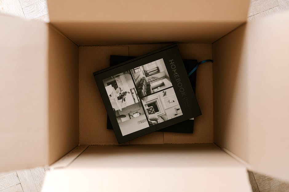 Inside a cardboard moving box viewed from above, containing a black and white household handbook with images of living room furniture, decorative items, and interior spaces on its cover. The box is situated on a light wooden floor, with no other objects visible. The handbook appears to be part of packing and planning materials used during a home relocation process, highlighting the importance of organized packing when arranging furniture transport or removals. This setup is typical in professional house removals, where packing materials such as cardboard boxes and printed guides are used by movers from Movers Hounslow to ensure a smooth and efficient moving process, especially when avoiding common mistakes during bookings in Hounslow.
