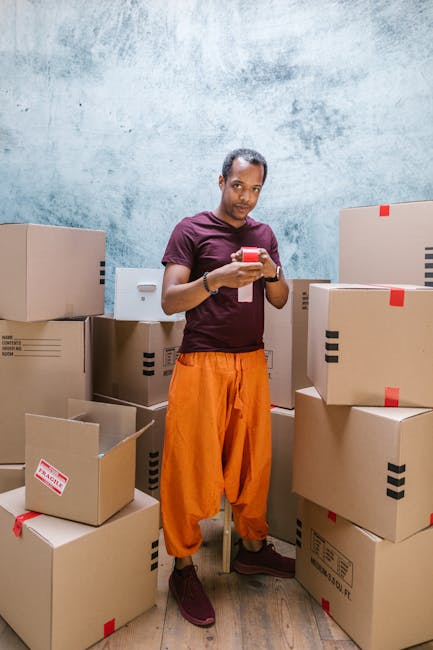A man standing inside a room surrounded by stacked cardboard boxes, some sealed with red tape and others open revealing packing materials. He is wearing a dark red T-shirt and bright orange loose trousers, holding and looking at a smartphone. The boxes vary in size and are arranged on the floor and on a small table, suggesting a packing or unpacking stage of a house removal process. The room has a textured light blue wall, and the lighting is neutral and evenly distributed. This scene illustrates the preparation phase of furniture transport and packing within a home relocation project, typical of professional removals services provided by Movers Hounslow.