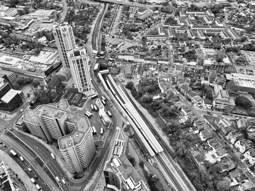 Aerial black-and-white photograph of a dense urban area showing a mix of high-rise buildings, mid-rise offices, and residential houses. Roads with visible traffic, including multiple buses and cars, r