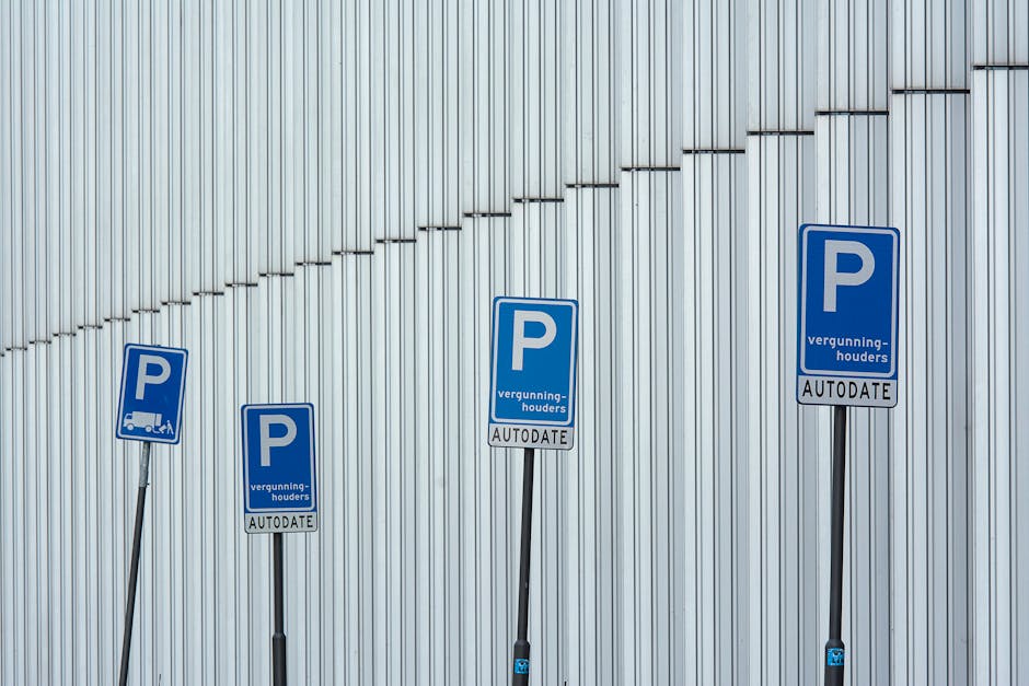 A wide, nearly empty parking lot with clearly marked white lines designating individual parking spaces, situated in an urban area under a bright blue sky with scattered white clouds. Several leafless
