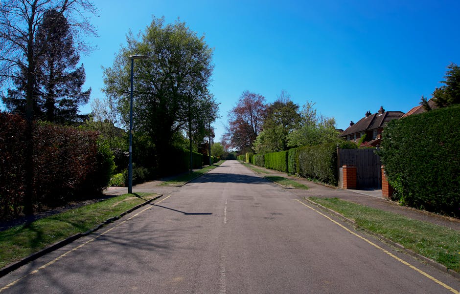 A quiet residential street in Hounslow with a clear blue sky overhead. On the left side, a tall lamppost casts a shadow on the pavement, while a narrow, paved road stretches into the distance, bordere