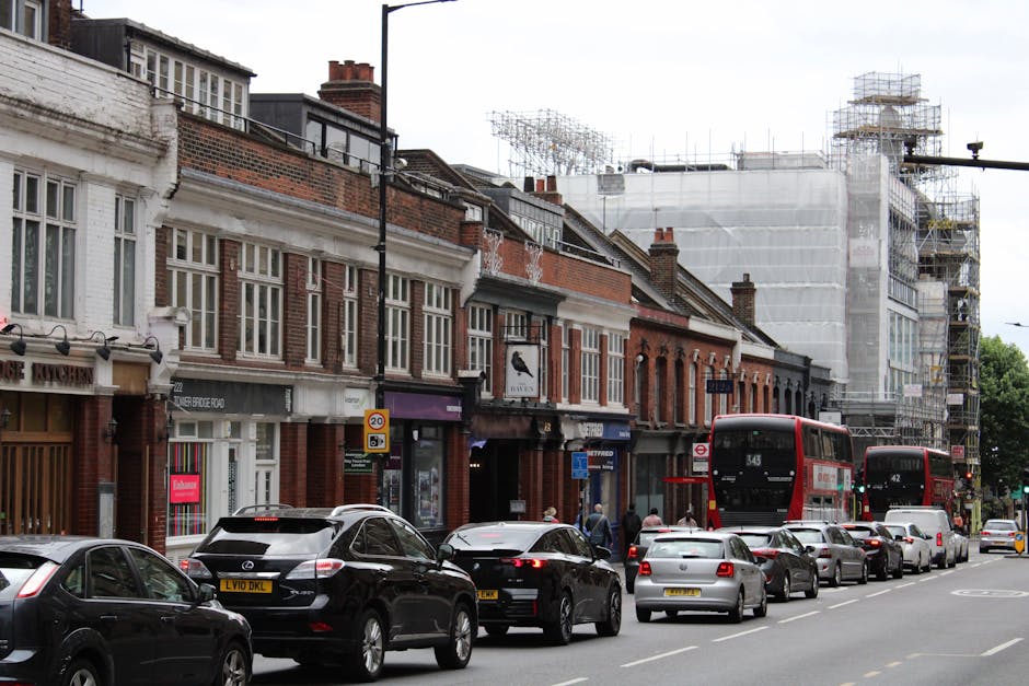 The image depicts a city street scene in Hounslow High Street TW3 during daytime, with a row of traditional brick andwood fronted premises, some of which are undergoing building work covered by scaffolding and white protective sheeting. parked along the curb are several vehicles including a black Mercedes-Benz estate, a silver hatchback, and a white van, with two red double-decker buses visible in the background, one with route number 343. Pedestrians are walking on the sidewalk near storefronts, which include retail shops and possibly cafes. The street is busy with parked cars and moving vehicles, and the overall scene captures a typical urban environment with ongoing building maintenance or refurbishment, consistent with local construction activity seen in Hounslow. This environment is suitable for illustrating themes of house removals or furniture transport, highlighting aspects of urban logistics and moving processes. The image emphasizes a mix of static parked vehicles, ongoing construction, and street-level activity fitting for a professional removals company like Movers Hounslow, specializing in residential relocations and moving logistics.