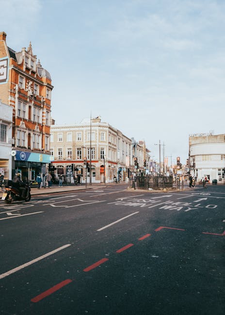 A street scene showing a busy intersection on Hounslow High Street TW3, with Victorian and modern-style buildings lining both sides. The buildings have detailed brickwork, large windows, and decorative facades. The sky is partly cloudy with soft lighting illuminating the scene. Pedestrians are walking along the pavement, and there are vehicles, including a motorcycle and a parked car, visible near the curb. Markings on the road indicate traffic directions and designated lanes for vehicles, with a dashed red line at the edge of the pavement. In the foreground, part of a loading area or pavement space is visible, suggesting a location where home relocation or furniture transport activities may take place. This setting is associated with moving services, such as those provided by Movers Hounslow, who specialise in house removals and furniture transport in the area.