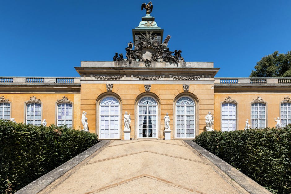 A wide exterior view of a historic building with a grand staircase leading up to three large arched windows, flanked by classical white statues positioned on pedestals along the stone balustrade. The building features a yellow facade with decorative stone accents and a prominent ornate sculpture at the top, set against a clear blue sky. In the foreground, a paved pathway descends from the entrance, bordered by well-maintained green hedges, suggesting a setting suitable for house removals or relocation services. The scene is well-lit by natural daylight, highlighting the architectural details and the peaceful environment, which aligns with planning for furniture transport and home relocation activities. Movers Hounslow, offering professional removals, may use this area for efficient loading and unloading processes associated with house removals and moving logistics near Syon Park.