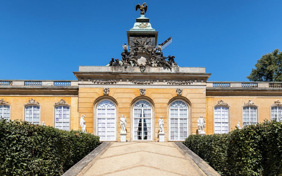 A wide exterior view of a historic building with a grand staircase leading up to three large arched windows, flanked by classical white statues positioned on pedestals along the stone balustrade. The building features a yellow facade with decorative stone accents and a prominent ornate sculpture at the top, set against a clear blue sky. In the foreground, a paved pathway descends from the entrance, bordered by well-maintained green hedges, suggesting a setting suitable for house removals or relocation services. The scene is well-lit by natural daylight, highlighting the architectural details and the peaceful environment, which aligns with planning for furniture transport and home relocation activities. Movers Hounslow, offering professional removals, may use this area for efficient loading and unloading processes associated with house removals and moving logistics near Syon Park.